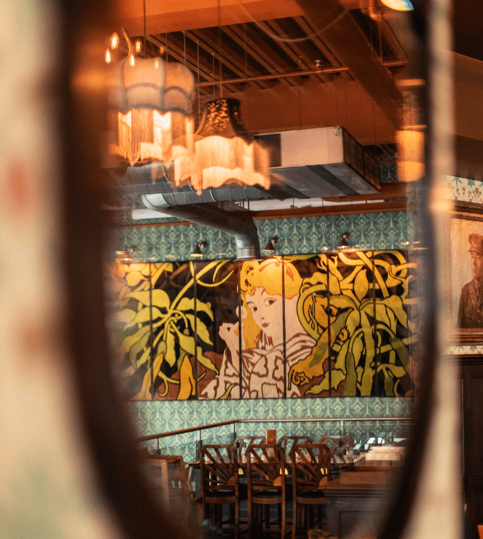 A cozy restaurant interior reflected in a mirror, featuring a mural of a woman with yellow hair surrounded by green leaves, with empty wooden chairs and tables beneath warm hanging lights.