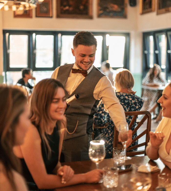 A smiling waiter in a vest and bow tie serves drinks to a group of women seated around a wooden table in a warmly lit restaurant. The atmosphere is lively and social, with people in the background.