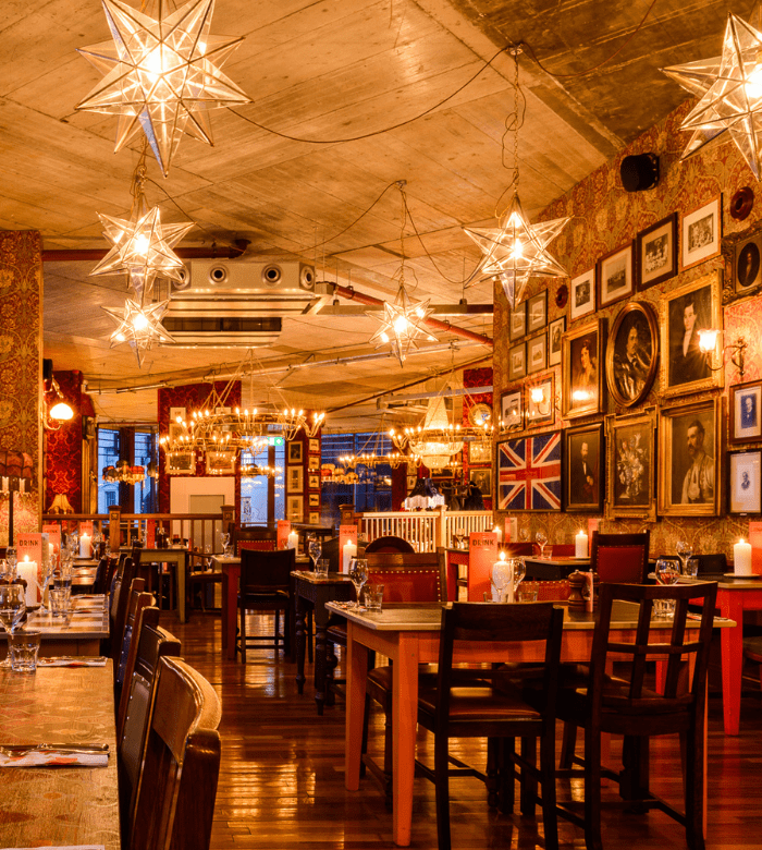 A warmly lit restaurant with star-shaped lights, wooden tables and chairs, candles, and walls decorated with portraits, framed art, and a British flag. The atmosphere is cozy and inviting.