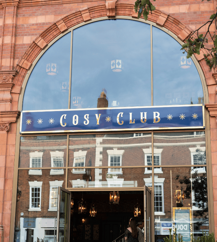 The entrance of Cosy Club, with a large arched window and a blue and gold sign, set in a brick building reflecting opposite buildings in the glass.