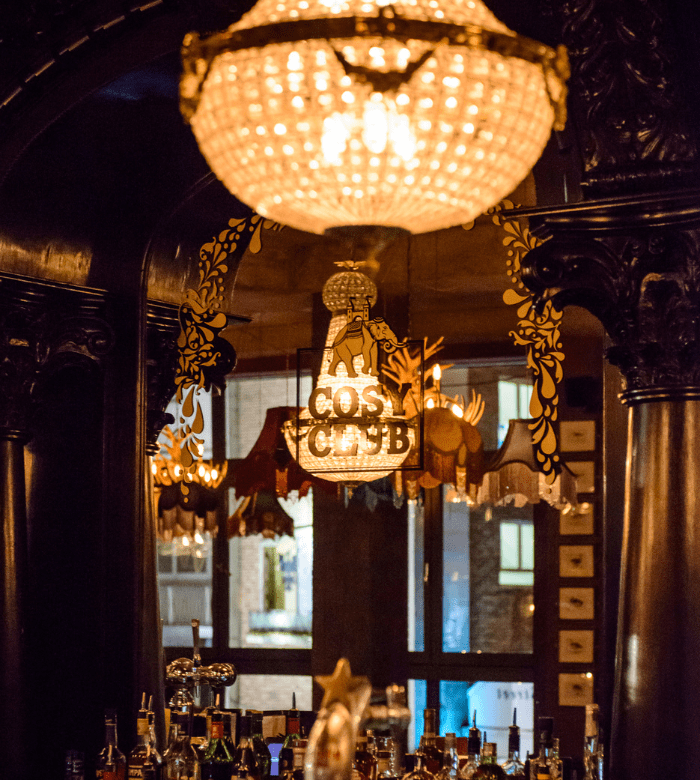 A chandelier hangs above an ornate bar with a mirror reflecting a decorative “Cosmic Club” sign, surrounded by dark wood columns and shelves of bottles, with warm lighting and windows in the background.