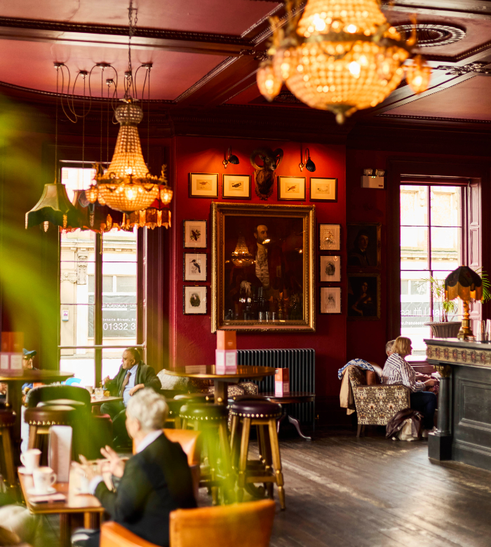 Cozy, vintage-style cafe with red walls, chandeliers, wooden tables, and eclectic wall art. People are seated, chatting and enjoying drinks by large windows letting in warm natural light.