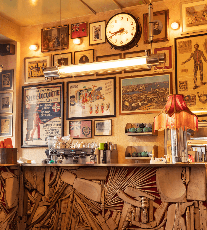A vintage café interior with eclectic wall art, a large clock, framed posters, and photos. The counter has a quirky wooden design, and a red-shaded lamp sits beside a coffee machine and stacked cups.