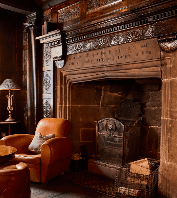 A cozy room with leather armchairs, a decorative stone fireplace with carved details, an ornate wooden mantel, a metal fireguard, a wire basket with firewood, and a classic lamp on a side table.