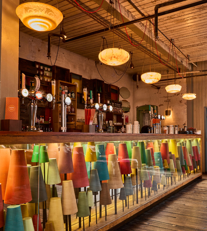 A cozy bar interior with warm lighting, colorful cone-shaped decorations under a glass counter, beer taps on the wooden bar, and shelves with glasses, bottles, and mugs in the background.