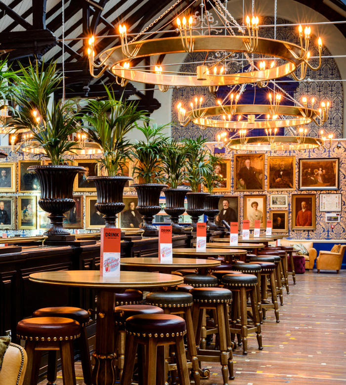 A stylish bar interior with round wooden tables, cushioned stools, large potted plants, and elegant chandeliers. Portrait paintings and blue patterned tile cover the back wall, creating a sophisticated, inviting atmosphere.