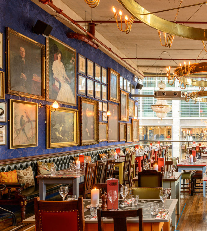 A stylish restaurant interior with eclectic framed art on blue walls, elegant chandeliers, set tables with glassware, cushioned seating, and large windows letting in natural light.