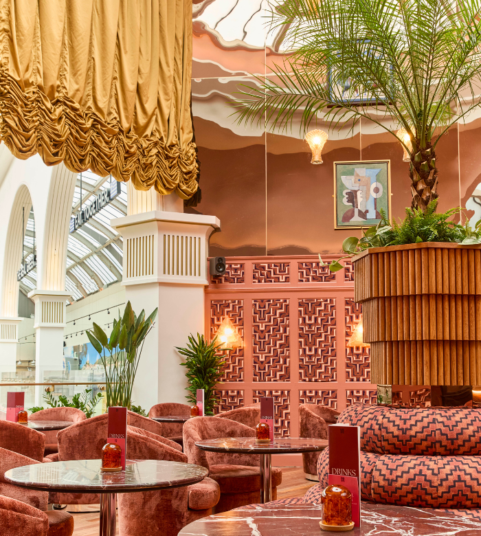 A stylish lounge with pink velvet chairs, marble tables, lush green plants, a large palm, ornate gold drapery, and geometric wall designs under a domed ceiling with soft lighting.