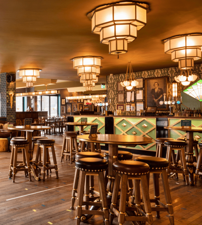 A spacious, warmly lit bar with wooden floors, round tables, cushioned stools, art deco chandeliers, and a stylish bar counter in the background, decorated with framed pictures and patterned wallpaper.