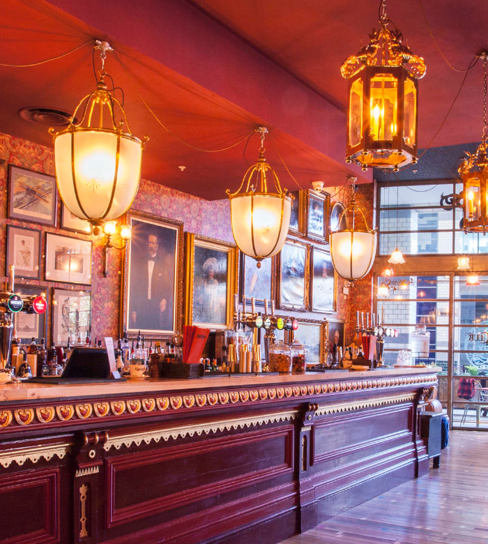 A vintage-style bar with ornate hanging lamps, framed pictures and portraits on patterned wallpaper, a long wooden counter, and large windows letting in natural light.