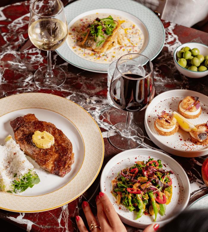 A marble table set with a steak topped with butter, a salad, deviled eggs, green olives, a white fish dish, a glass of red wine, and a glass of white wine. A hand with red nail polish is visible in the foreground.