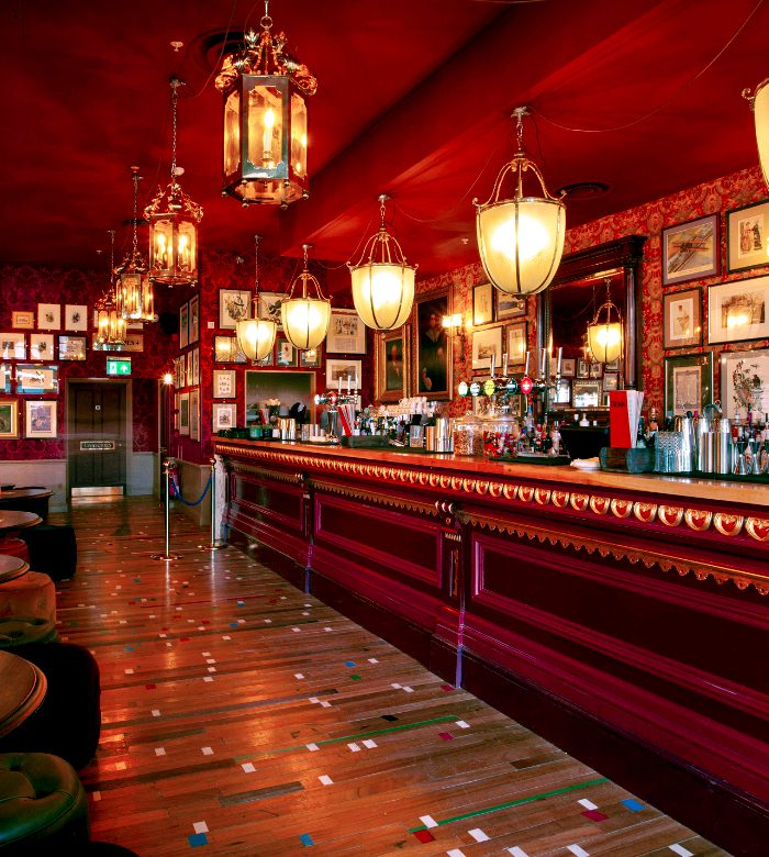 A stylish bar with ornate red decor, large lantern-style lights, framed pictures on the walls, a polished wooden floor, and a long bar counter with assorted bottles and glassware.
