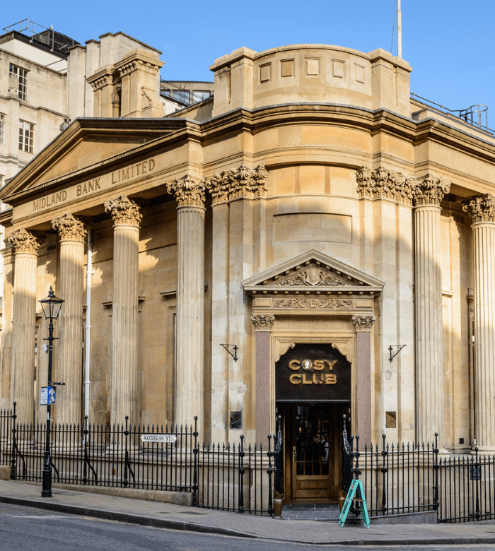 A historic neoclassical building with tall columns and ornate details houses the Cosy Club restaurant, with "Midland Bank Limited" inscribed above the entrance. Black railings and a streetlamp line the sidewalk outside.