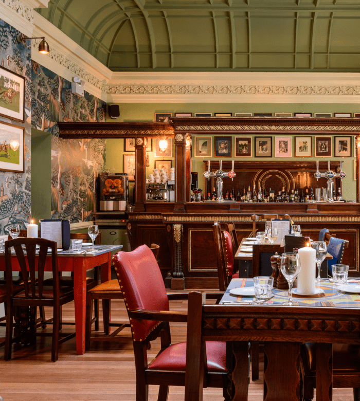 A cozy restaurant with patterned wallpaper, ornate green ceiling, wooden furniture, and tables set with candles and glasses. A classic wooden bar with taps and framed artwork is in the background.