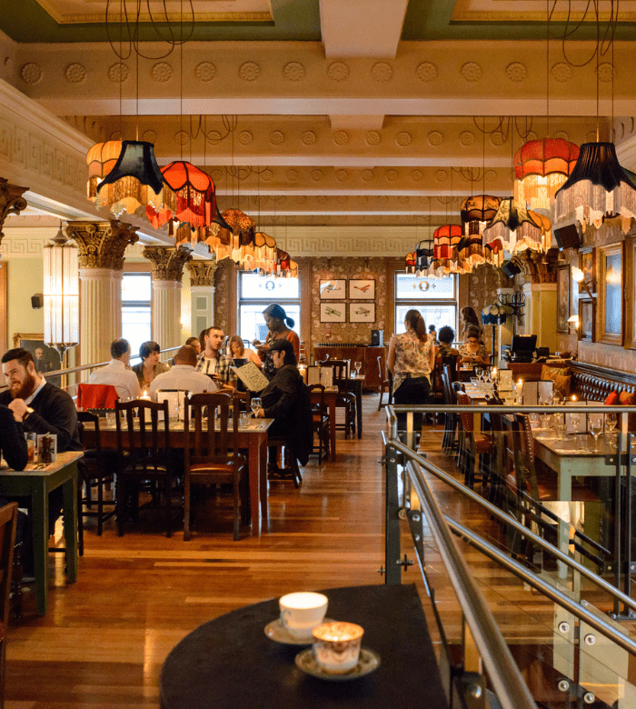 A cozy, warmly lit restaurant with vintage lamps hanging from the ceiling, wooden tables and chairs, people dining and chatting, and classic decor with ornate columns and framed pictures on the walls.