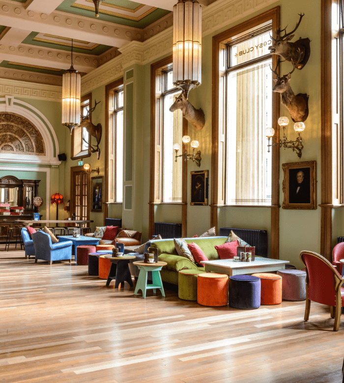 A spacious, elegant lounge with tall windows, colorful sofas and stools, wooden floors, high ceilings, hanging lights, wall-mounted deer heads, and framed portraits, creating a cozy yet sophisticated atmosphere.