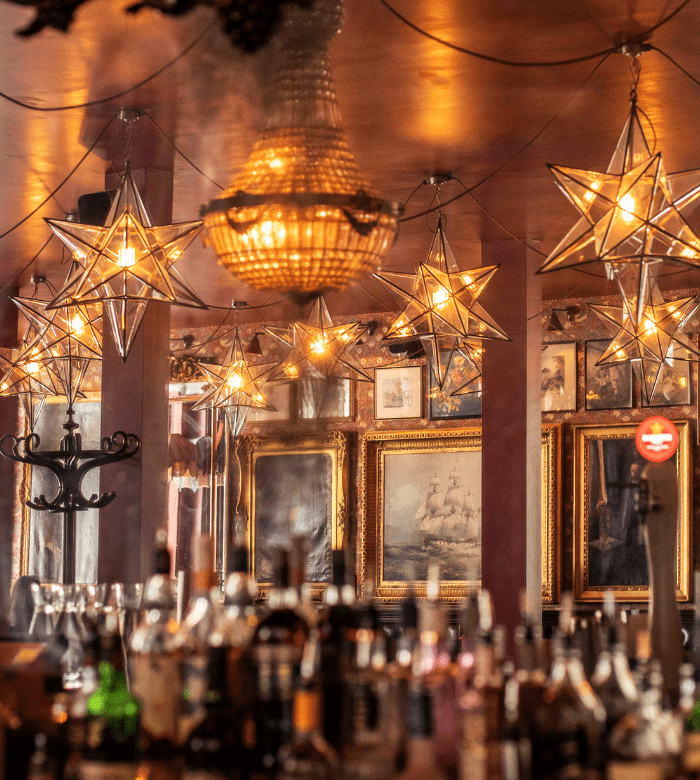 A warmly lit bar with bottles in the foreground, illuminated by star-shaped pendant lights and a crystal chandelier. Framed paintings and mirrors decorate the walls, creating a cozy, vintage ambiance.