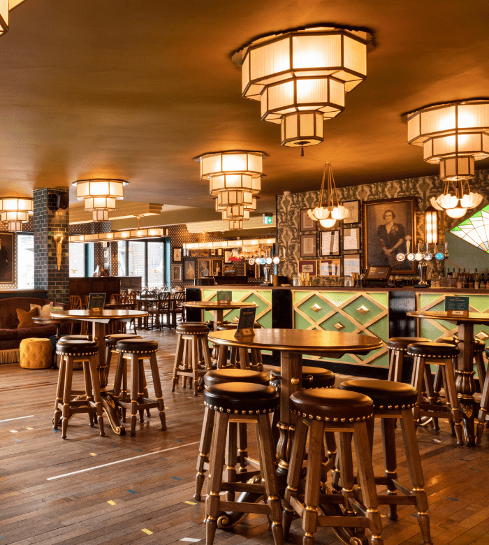 A stylish bar with Art Deco-inspired lighting, wooden stools around high tables, patterned wallpaper, and a long bar counter in the background, creating a warm, inviting atmosphere.