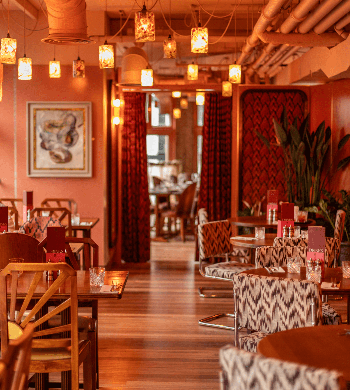A stylish restaurant interior with warm lighting, patterned chairs, wooden tables, red menus, and contemporary decor. Hanging pendant lights and plants add to the cozy, inviting atmosphere.