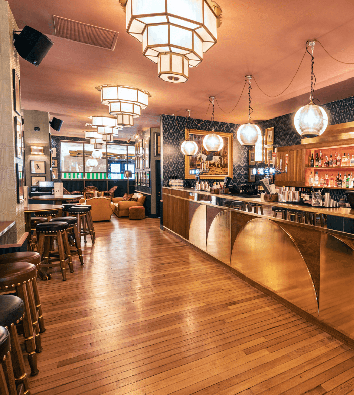 A stylish bar interior with wooden floors, gold accents, round bar stools, and plush armchairs. Large geometric and globe pendant lights hang from the ceiling, creating a warm, inviting atmosphere. The bar shelves are stocked with bottles.