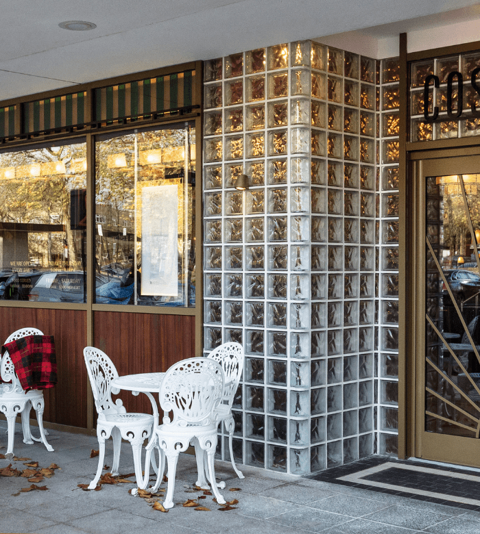 A small outdoor cafe area with two white ornate metal chairs and a table. One chair has a red plaid jacket draped over it. The cafe has glass block walls and large windows. Fallen leaves are scattered on the ground.