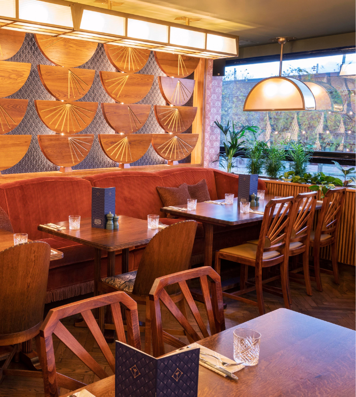 A cozy restaurant interior with wooden tables and chairs, orange cushioned seating, decorative fan-shaped wall art, potted plants by the window, and menus and glasses set on the tables.