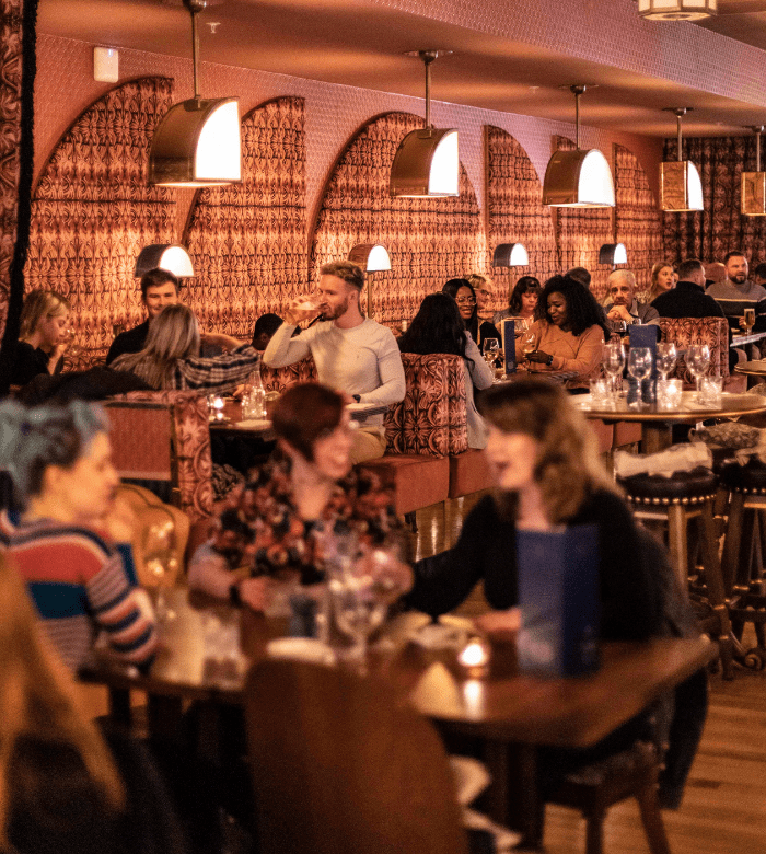 A lively restaurant with warm lighting, patterned booths, and people socializing at tables, enjoying food and drinks. The atmosphere appears cozy and vibrant, with groups engaged in conversation.