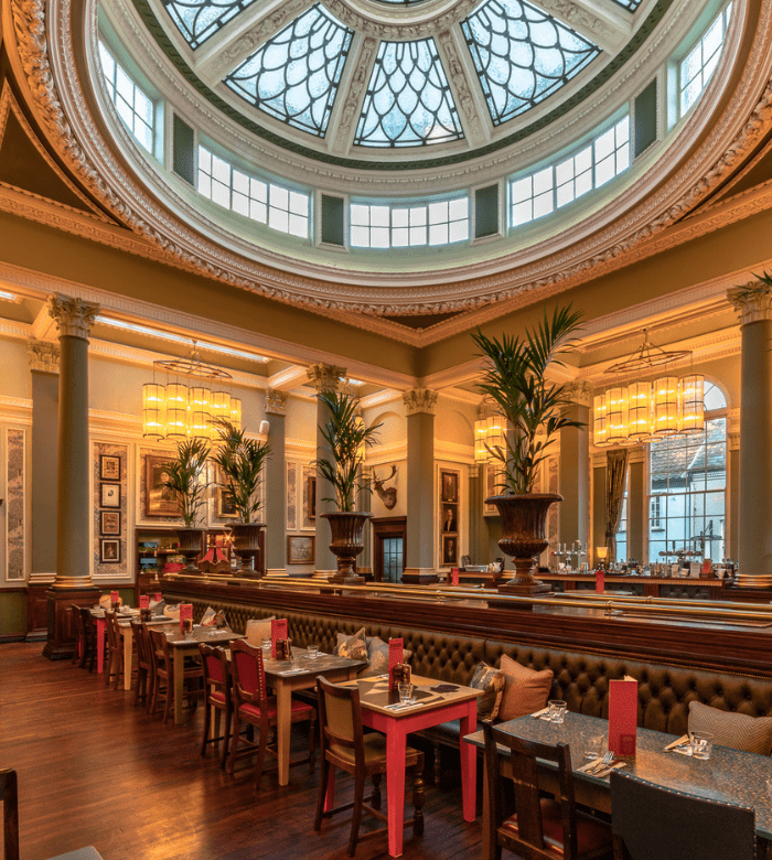 Elegant restaurant interior with a high glass dome ceiling, grand columns, large potted plants, wooden floors, and tables set for dining, creating a sophisticated and inviting atmosphere.