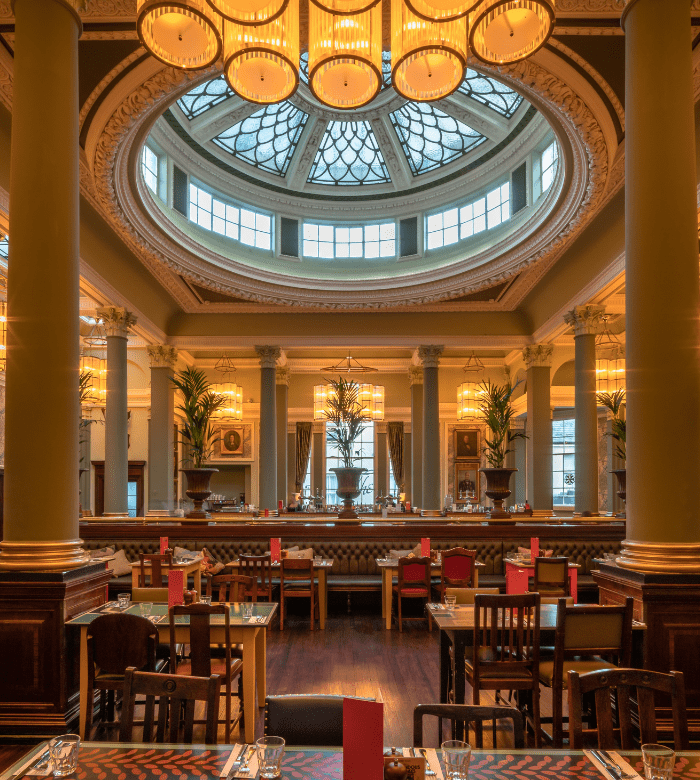 Elegant restaurant interior with high ceilings, large columns, and a grand glass dome skylight. Tables are set for dining, surrounded by ornate decor, warm lighting, and tall potted plants by the windows.