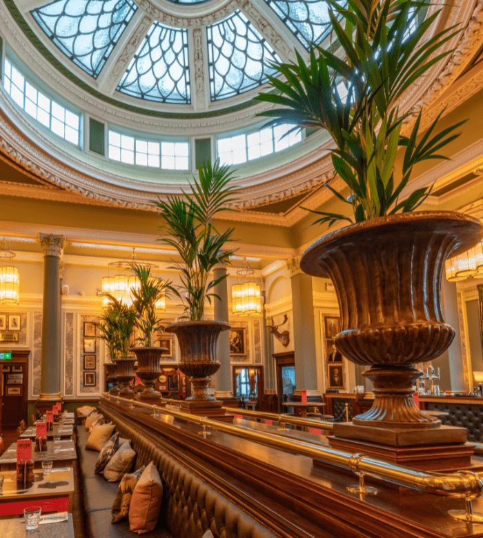 A grand restaurant interior with large potted plants, plush booth seating, elegant wood paneling, and a domed glass ceiling letting in natural light. The décor is classic and opulent, with framed art on the walls.