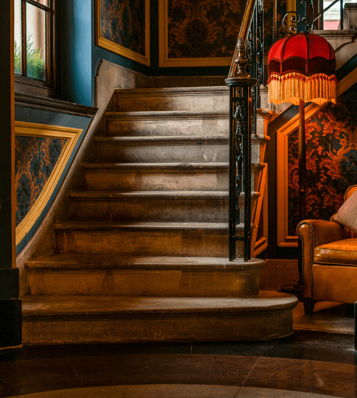 A vintage stone staircase with ornate railings beside a brown leather armchair and a fringed red lamp in a richly decorated, warmly lit room.