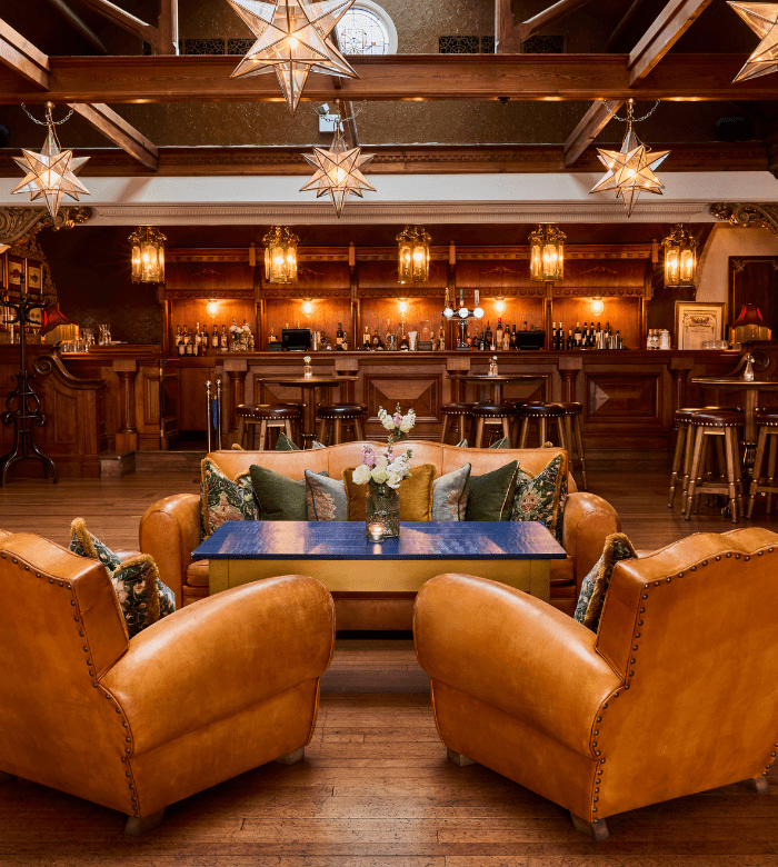 A cozy lounge with four tan leather armchairs around a wooden coffee table, floral centerpiece, and decorative pillows. Star-shaped pendant lights hang above, with a wooden bar and vintage stools in the background.