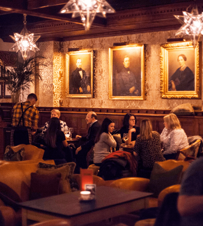 A cozy, warmly lit lounge with people sitting and chatting at tables. Ornate star-shaped lights hang from the ceiling, and three large vintage portraits are displayed on the wall behind them.