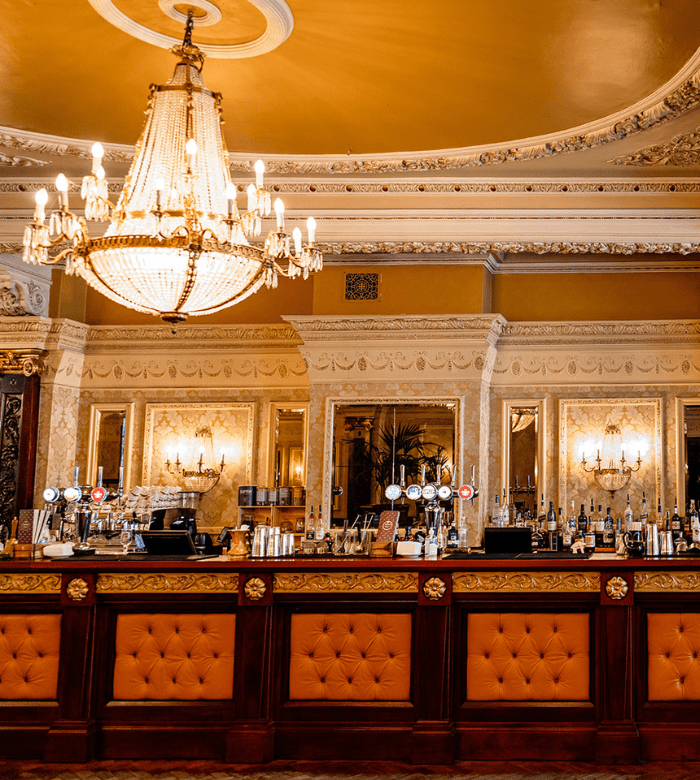 An ornate bar with a tufted orange front, crystal chandelier, and gold-trimmed mirrors and walls, creating an elegant and luxurious atmosphere. Various bottles and taps line the bar counter.