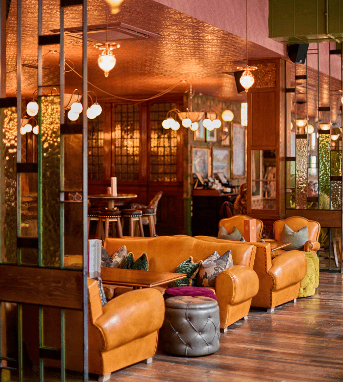 A cozy lounge with tan leather sofas, colorful cushions, a round ottoman, wooden floors, pendant lights, and a dining area in the background with warm, ambient lighting.