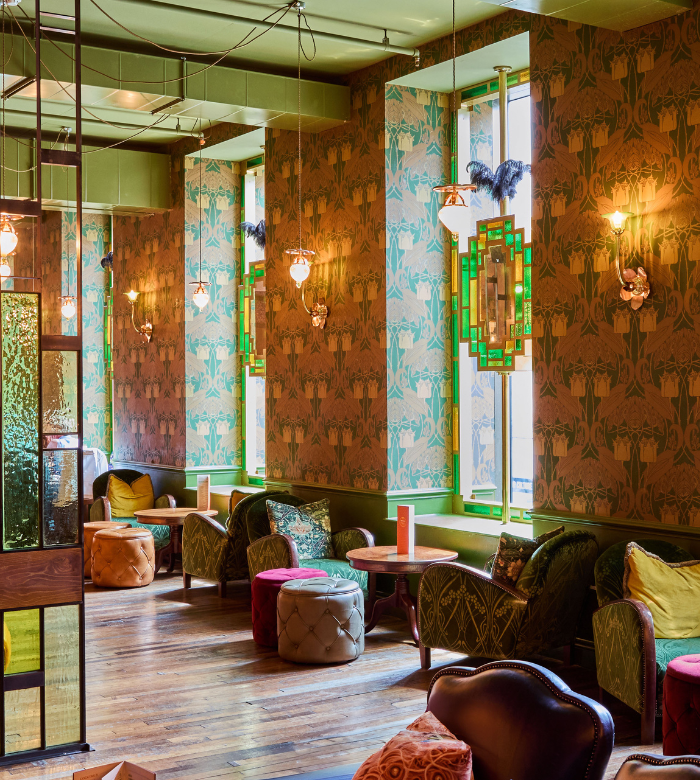 A cozy lounge with patterned wallpaper, vintage green and gold decor, plush chairs, ottomans, and hanging pendant lights, with natural light streaming through tall windows.