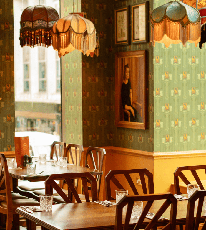 Cozy restaurant interior with wooden tables and chairs, vintage-style lamps, green patterned wallpaper, framed art, and set tables with glasses and cutlery near a window.