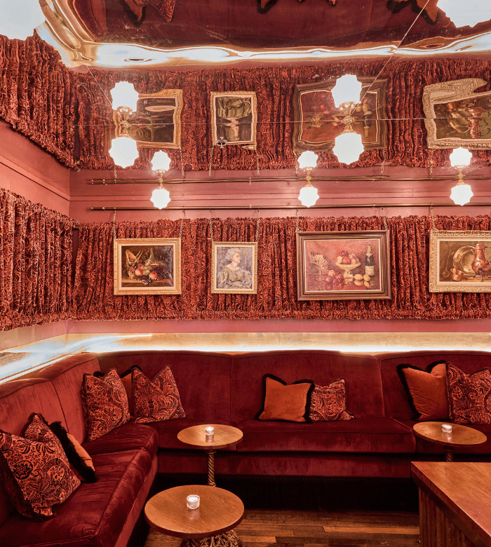 A cozy lounge with plush red velvet seating, patterned pillows, small round tables, ornate framed paintings on textured red walls, and warm pendant lights reflected in a mirrored ceiling.