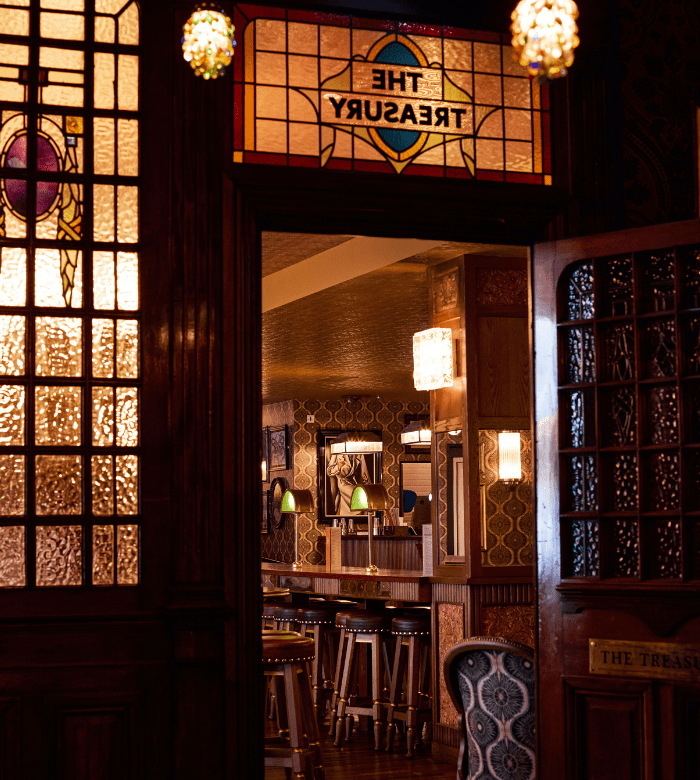Warmly lit pub interior seen through stained glass doors, with high stools, wooden tables, ornate wallpaper, and a sign reading "THE TREASURY" in stained glass above the entrance.