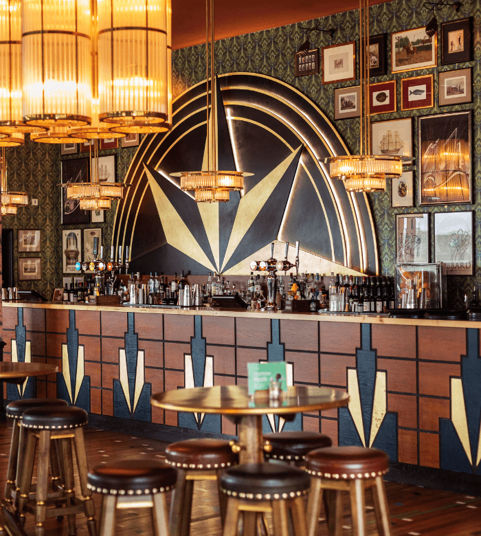 A stylish bar with an Art Deco design features geometric patterns, gold accents, vintage wallpaper, framed pictures on the wall, warmly lit chandeliers, and round wooden tables with stools in the foreground.