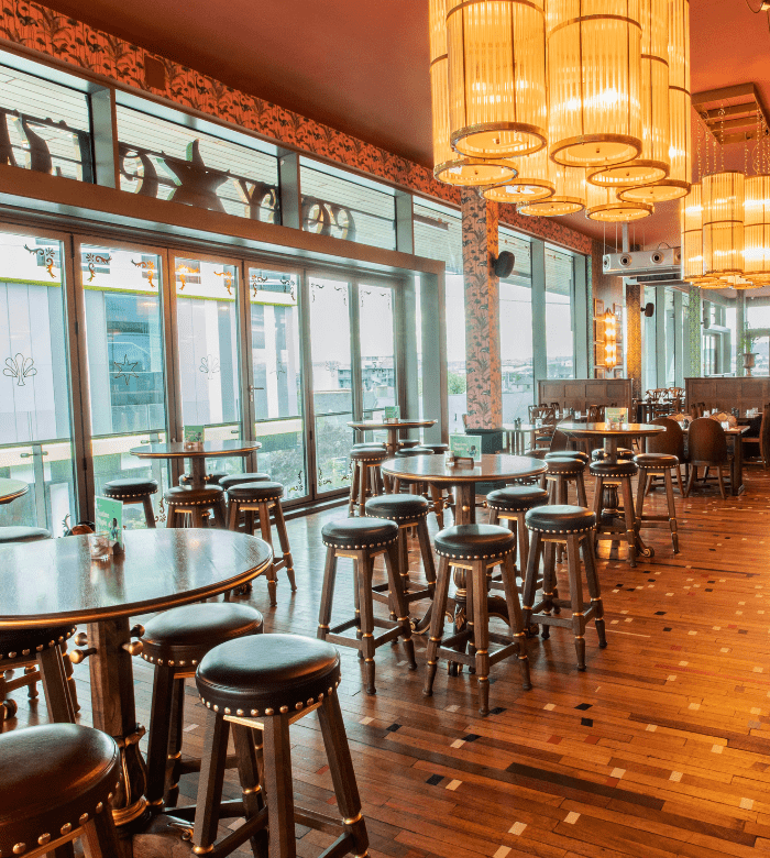 A stylish bar with round tables and wooden stools, large windows letting in natural light, patterned wallpaper, and multiple ornate pendant lights hanging from the ceiling.