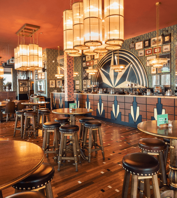 Art Deco-style bar with geometric patterns, wooden tables and stools, ornate chandeliers, and a mirrored wall behind the bar, decorated with framed photos and patterned wallpaper. Warm lighting creates a cozy, elegant atmosphere.