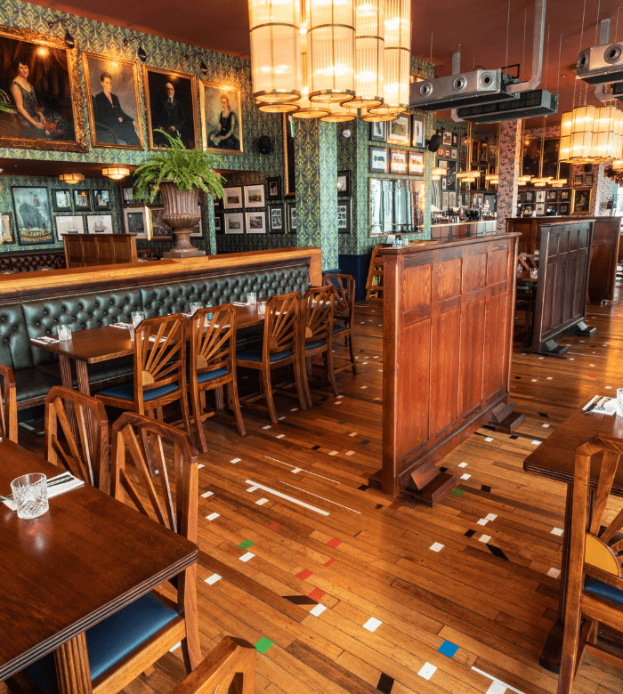 A stylish restaurant interior with wooden tables and chairs, green upholstered booths, vintage portraits and framed art on patterned walls, hanging pendant lights, and a hardwood floor with colorful geometric inlays.