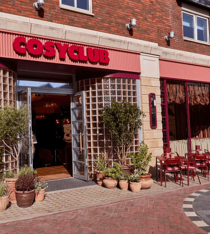 The entrance of a restaurant called Cosy Club, with open glass doors, potted plants near the doorway, and several red tables and chairs arranged outside on a brick pavement.