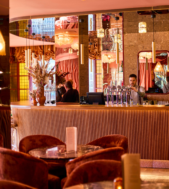 A bartender stands behind a stylish, curved bar with gold accents and taps, surrounded by plush, reddish chairs and marble tables in a warmly lit, elegant lounge with decorative mirrors and soft, vintage lighting.