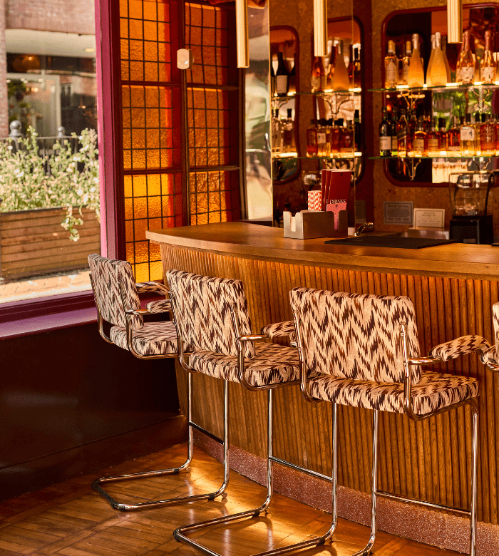 A stylish bar interior with patterned upholstered chairs, a wooden counter, hanging lights, and shelves of bottles. Large windows let in sunlight, revealing greenery and an outdoor area.