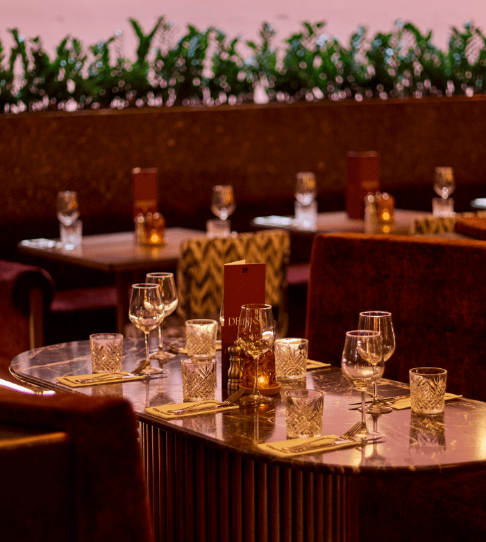 Elegant restaurant table set with wine glasses, water glasses, cutlery, and menus. The background shows more tables and green plants along a ledge, creating a warm, inviting atmosphere.
