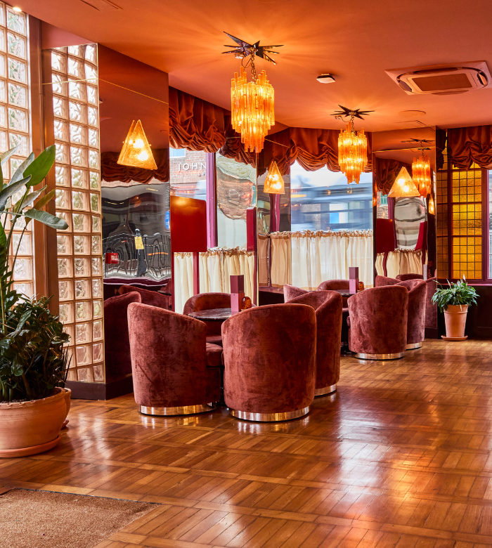 Cozy lounge with plush red velvet chairs, round tables, and warm wood floors. Elegant chandeliers hang from the ceiling. Large potted plants and glass block walls add to the retro, inviting atmosphere.