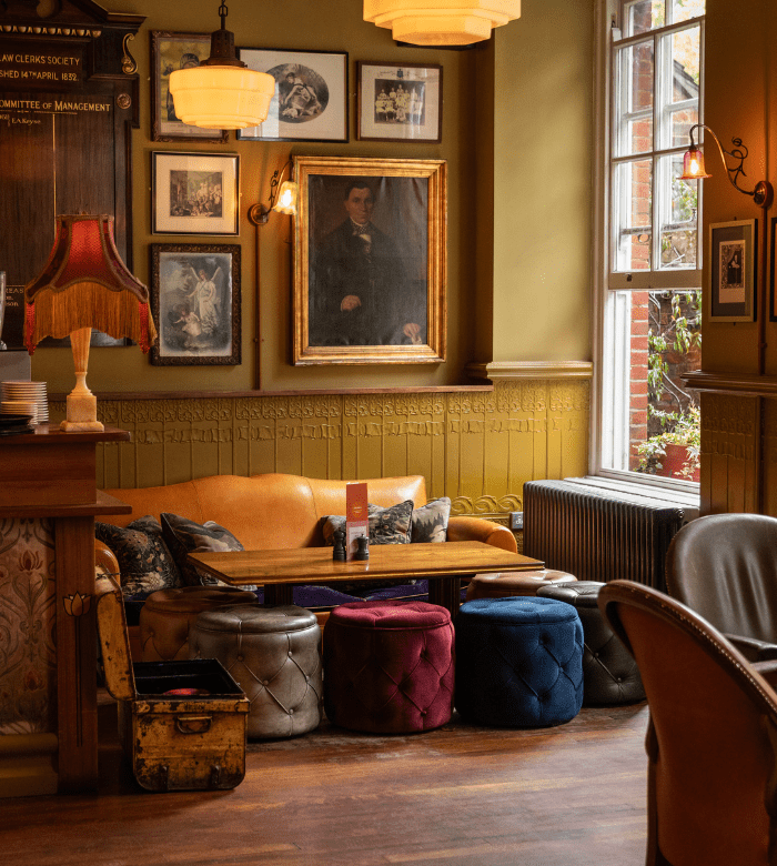 A cozy lounge area with a tan leather sofa, velvet ottomans in various colors, a wooden table, vintage lamps, framed art on the walls, and sunlight streaming through a large window.