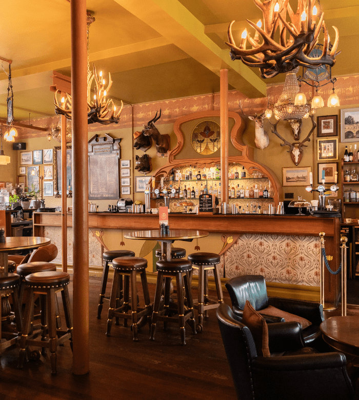 A cozy, vintage-style bar with high stools, wooden tables, animal head mounts on patterned walls, chandeliers, and a well-stocked bar counter in a warmly lit, rustic interior.